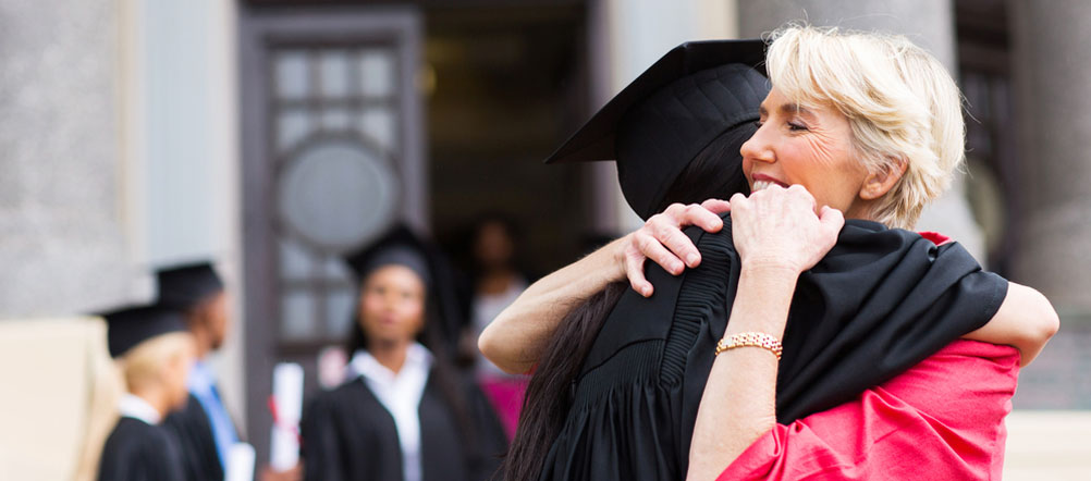 A young graduate, wearing a cap and gown, is hugging a lady with other graduates in the background.
